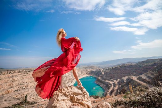 Side view of a beautiful sensual woman in a red long dress posing on a rock high above the lake in the afternoon. Against the background of the blue sky and the lake in the form of a heart.