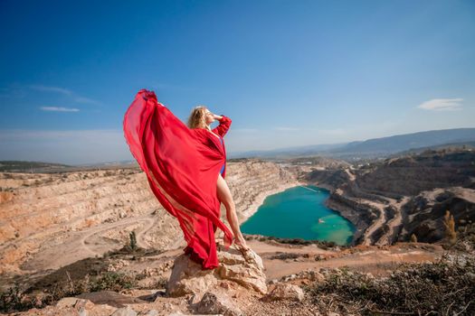 Side view of a beautiful sensual woman in a red long dress posing on a rock high above the lake in the afternoon. Against the background of the blue sky and the lake in the form of a heart.