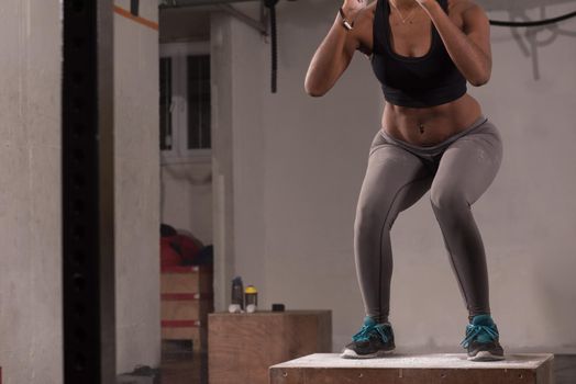 Fit young african american woman box jumping at a crossfit style gym. Female athlete is performing box jumps at gym with focus on legs