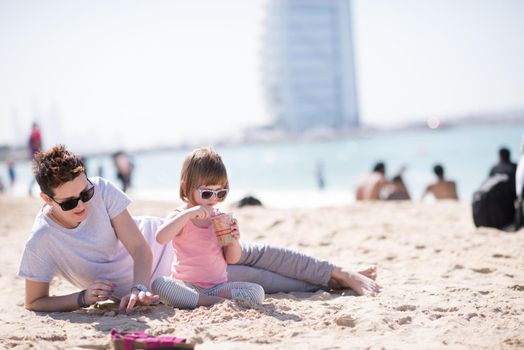 cool young mom and her little girl playing with a bucket full of sand wearing sunglasses and relaxing on the beach on a sunny day