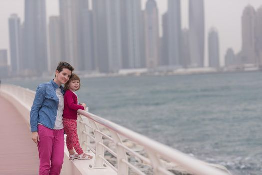 young mother and cute little girl running and cheerfully spend their time on the promenade by the sea with a big city in the background