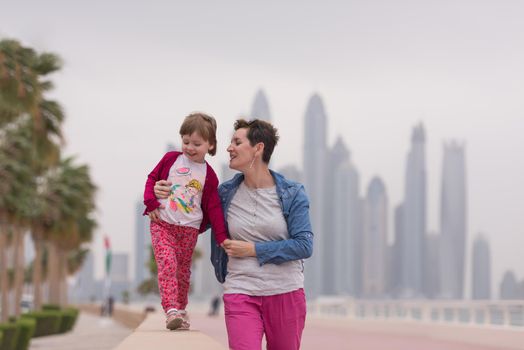 young mother and cute little girl running and cheerfully spend their time on the promenade by the sea with a big city in the background