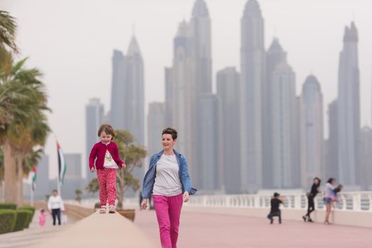 young mother and cute little girl running and cheerfully spend their time on the promenade by the sea with a big city in the background