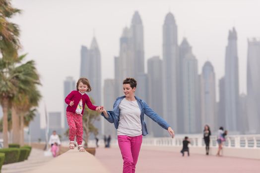 young mother and cute little girl running and cheerfully spend their time on the promenade by the sea with a big city in the background