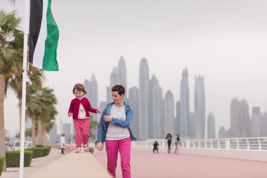 young mother and cute little girl running and cheerfully spend their time on the promenade by the sea with a big city in the background