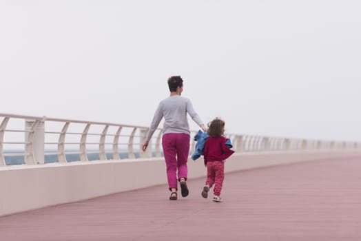 young mother and cute little girl running and cheerfully spend their time on the promenade by the sea