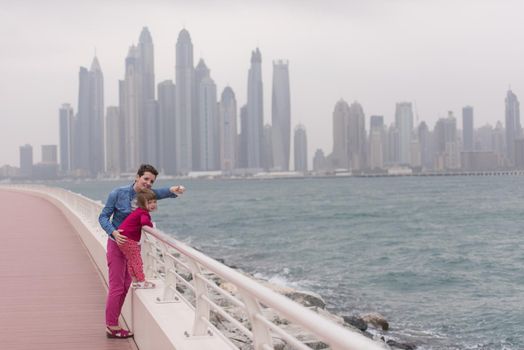 young mother and cute little girl running and cheerfully spend their time on the promenade by the sea with a big city in the background