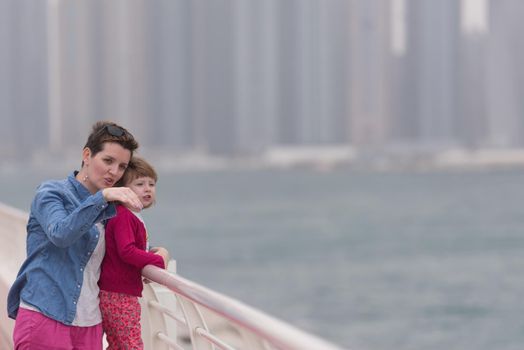 young mother and cute little girl running and cheerfully spend their time on the promenade by the sea with a big city in the background