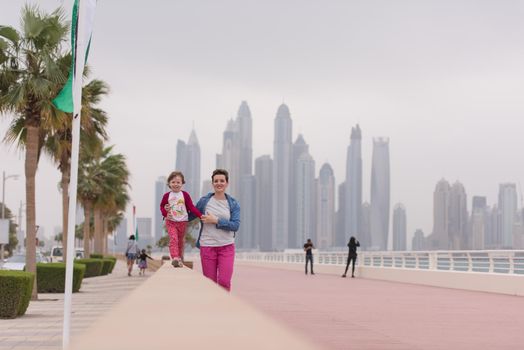 young mother and cute little girl running and cheerfully spend their time on the promenade by the sea with a big city in the background