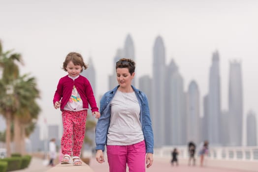 young mother and cute little girl running and cheerfully spend their time on the promenade by the sea with a big city in the background