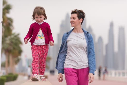 young mother and cute little girl running and cheerfully spend their time on the promenade by the sea with a big city in the background