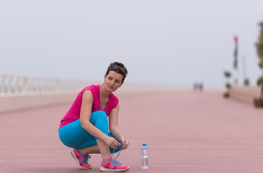 Young woman tying shoelaces on sneakers on a promenade. Standing next to a bottle of water. Exercise outdoors