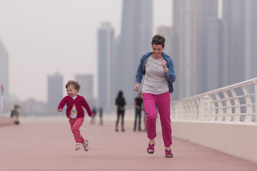 young mother and cute little girl running and cheerfully spend their time on the promenade by the sea with a big city in the background