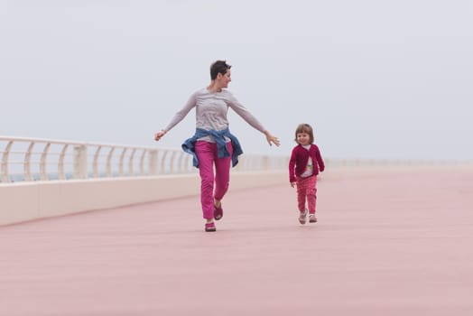 young mother and cute little girl running and cheerfully spend their time on the promenade by the sea