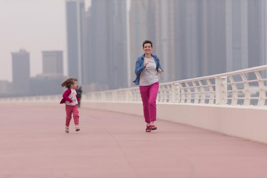 young mother and cute little girl running and cheerfully spend their time on the promenade by the sea with a big city in the background