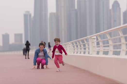 young mother and cute little girl running and cheerfully spend their time on the promenade by the sea with a big city in the background