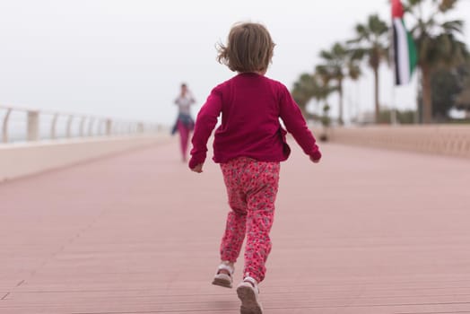 young mother and cute little girl running and cheerfully spend their time on the promenade by the sea