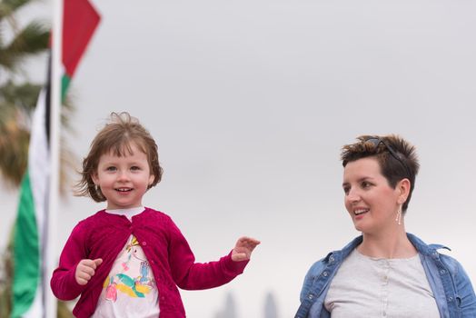 young mother and cute little girl running and cheerfully spend their time on the promenade by the sea with a big city in the background