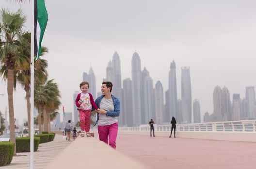young mother and cute little girl running and cheerfully spend their time on the promenade by the sea with a big city in the background