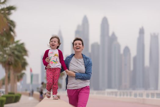 young mother and cute little girl running and cheerfully spend their time on the promenade by the sea with a big city in the background