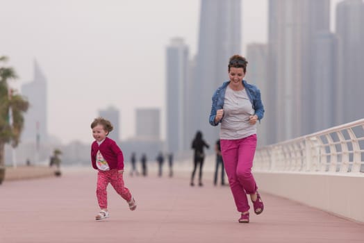 young mother and cute little girl running and cheerfully spend their time on the promenade by the sea with a big city in the background