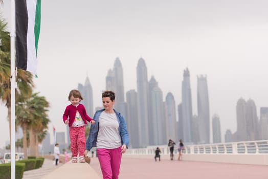 young mother and cute little girl running and cheerfully spend their time on the promenade by the sea with a big city in the background