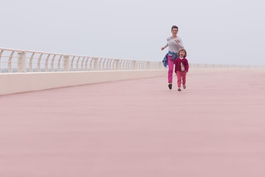 young mother and cute little girl running and cheerfully spend their time on the promenade by the sea