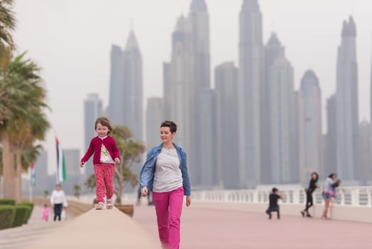 young mother and cute little girl running and cheerfully spend their time on the promenade by the sea with a big city in the background