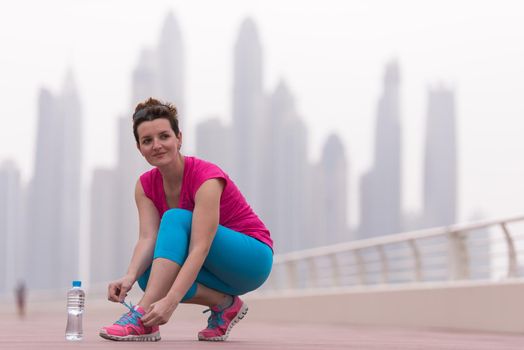 Young woman tying shoelaces on sneakers on a promenade with a big city in the background. Standing next to a bottle of water. Exercise outdoors