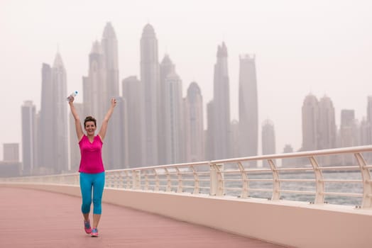 young woman celebrating a successful training run on the promenade by the sea with a bottle of water and her hands raised in the air with a big city in the background