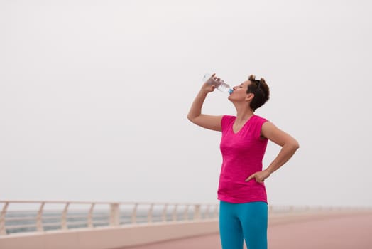 Fitness woman drinking water after running at the promenade by the sea. Thirsty sport runner resting taking a break with water bottle drink outside after training. Beautiful fit sporty girl.