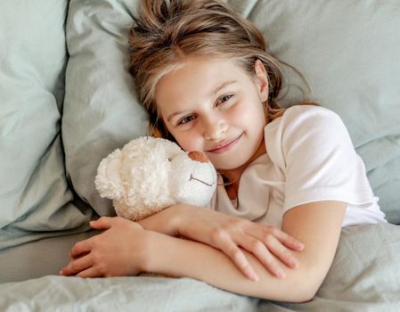 Cute child girl hugging teddy bear toy in the bed and smiling. Pretty kid in morning in bedchamber