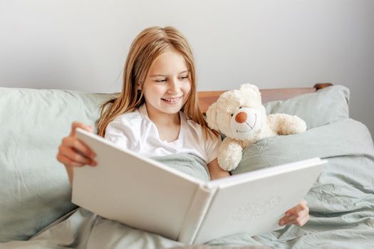 Beautiful child girl with teddy bear toy in the bed reading book. Cute female kid resting in bedchamber