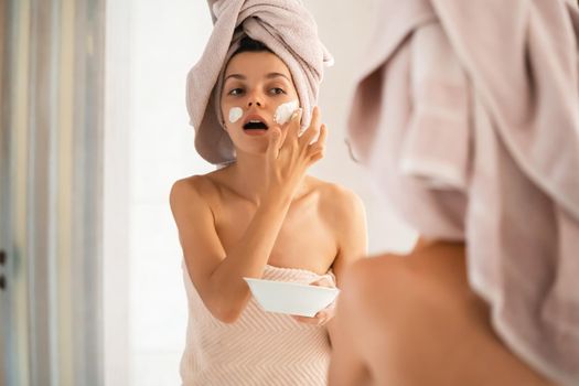 A young girl in a towel on her head and body stands near the mirror in the bathroom and applies a clay mask to her face, the woman takes care of her health and beauty.