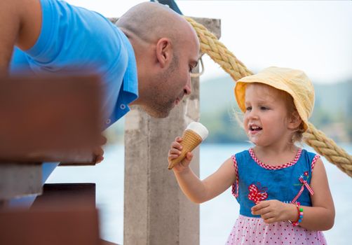 cute little girl having fun with her young father while eating ice cream by the sea during summer vacation holidays, celebration, children and people concept