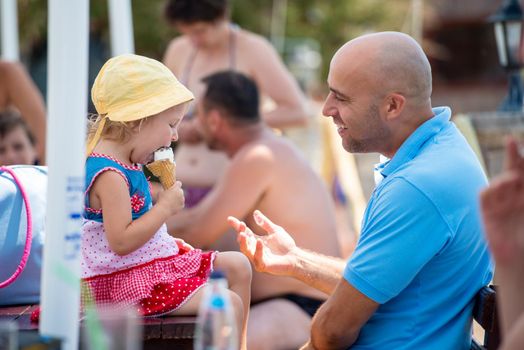 cute little girl having fun with her young father while eating ice cream by the sea during summer vacation holidays, celebration, children and people concept