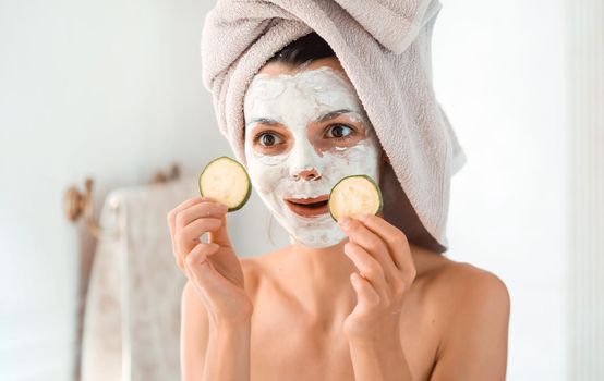 A young amusing girl in a white clay mask on her face, with a towel on her head, makes grimaces in front of the mirror and puts cucumber slices over her eyes.