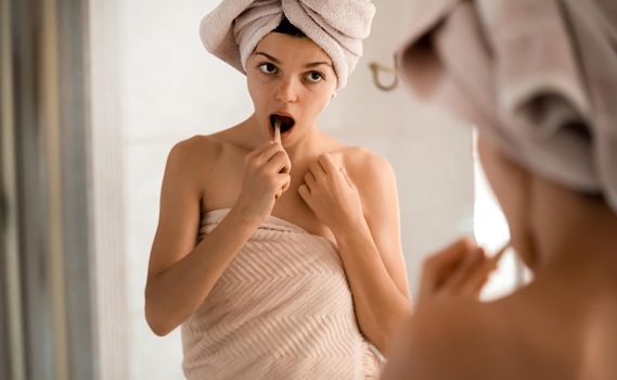 A young girl brushes her teeth near the mirror after relaxing in the bathroom, the woman is wrapped in a towel and takes care of the health and beauty of her body and teeth, gums.