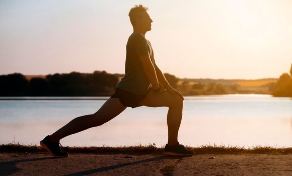 A young man trains outdoors against the backdrop of a beautiful lake at sunset, the athlete warms up, does exercises on the legs before running.