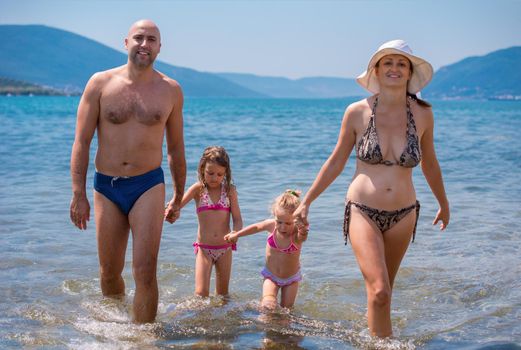 portrait of a young happy couple with cute little daughters having fun while standing in the shallow water of the sea during Summer vacation  Healthy family holiday concept