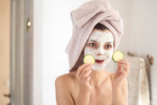 A young amusing girl in a white clay mask on her face, with a towel on her head, makes grimaces in front of the mirror and puts cucumber slices over her eyes.