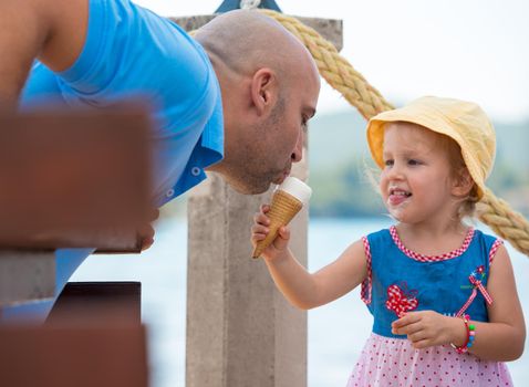 cute little girl having fun with her young father while eating ice cream by the sea during summer vacation holidays, celebration, children and people concept