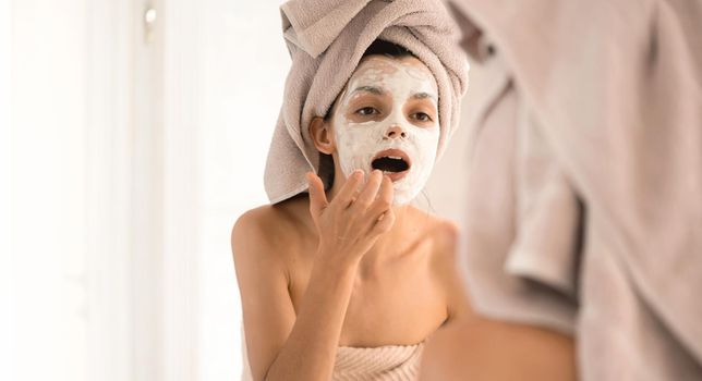 A young girl in a towel on her head and body stands near the mirror in the bathroom and applies a clay mask to her face, the woman takes care of her health and beauty.