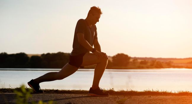 A young man trains outdoors against the backdrop of a beautiful lake at sunset, the athlete warms up, does exercises on the legs before running.