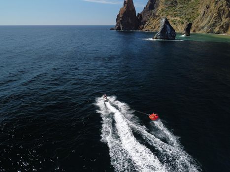 Happy people swim on air mattress behind a high speed water bike. Tourists ride on inflatable watercraft boat. Sea attraction. Water boat skating people on large Inflatable raft. Aerial copter view