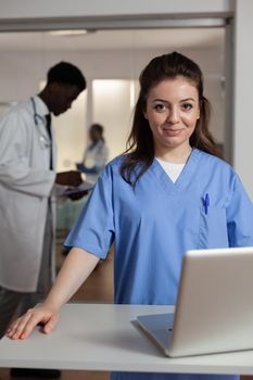 Portrait of smiling specialist therapist assistant woman standing at desk in hospital ward working at healthcare treatment typing medical expertise. In background clinical team monitoring sick patient