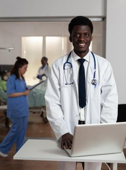 Portrait of smiling african american specialist therapist doctor typing sickness expertise on computer working at healthcare treatment in hospital ward. Medical team supporting patient after surgery