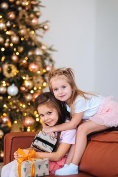 Two little girls are playing with New Year's gifts by the tree at sofa. Christmas, New Year tree.