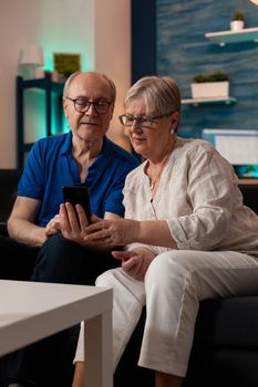 Retired husband and wife looking at smartphone screen in living room. Caucasian senior couple using digital device and technology while sitting on couch. Aged people enjoying retirement