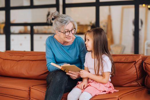Nice elderly woman grandmother reading story to granddaughter. Happy family at home concept